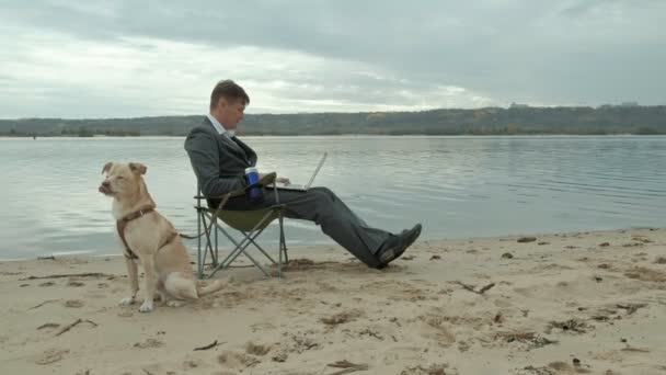 Mature businessman with dog sitting and relaxing near a river. Man in suit and using laptop. — Stock Video