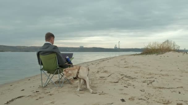 Reifer Geschäftsmann mit Hund, der in der Nähe eines Flusses sitzt und relaxt. Mann im Anzug und mit Laptop. — Stockvideo
