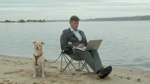 Rijpe zakenman met hond zitten en ontspannen in de buurt van een rivier. Man in pak en met behulp van laptop. — Stockvideo