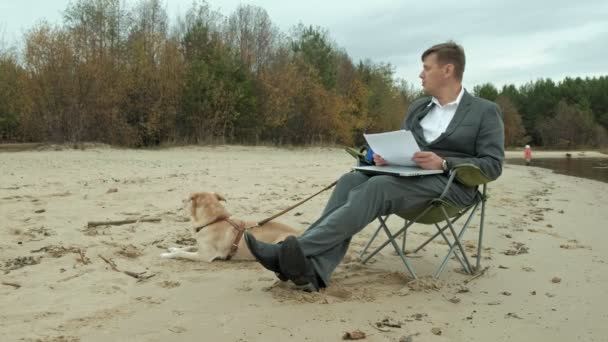 Starší podnikatel se psem, posezení a relaxaci u řeky. Muž v obleku a s pomocí přenosného počítače. — Stock video