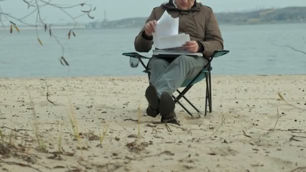 Ältere Geschäftsleute sitzen und entspannen in der Nähe eines Flusses. Mann in warmer Jacke und mit Laptop. — Stockvideo