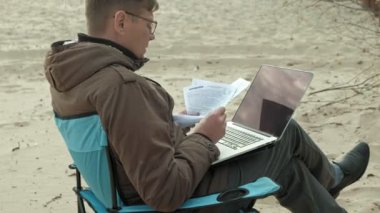 Oturma ve bir nehir kenarında rahatlatıcı olgun iş adamı. Adam sıcak bir ceket ve dizüstü bilgisayar kullanarak.