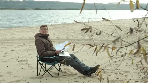 成熟的商人坐在河边放松。穿着保暖夹克的人, 使用笔记本电脑. — 图库视频影像