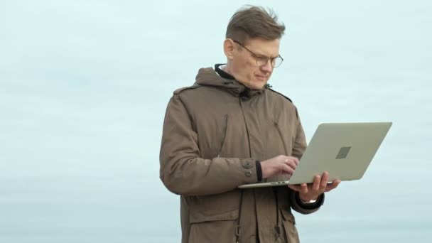 Rijpe zakenman zitten en ontspannen in de buurt van een rivier. Man in een warme jas en met behulp van laptop. — Stockvideo