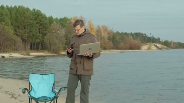 Ältere Geschäftsleute sitzen und entspannen in der Nähe eines Flusses. Mann in warmer Jacke und mit Laptop. — Stockvideo