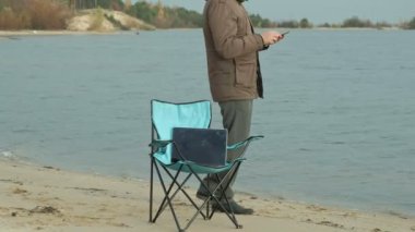 Oturma ve bir nehir kenarında rahatlatıcı olgun iş adamı. Adam sıcak bir ceket ve bir smartphone kullanarak.