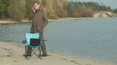 Oturma ve bir nehir kenarında rahatlatıcı olgun iş adamı. Adam sıcak bir ceket ve bir smartphone kullanarak.