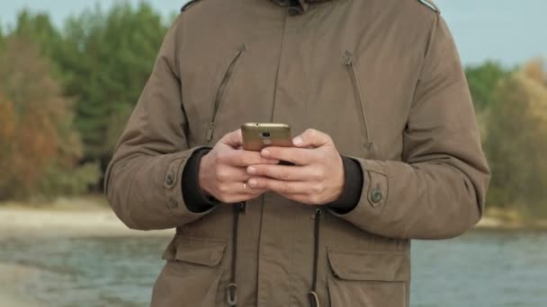 Mogen affärsman sitter och avkopplande nära en flod. Man i en varm jacka och med en smartphone. — Stockvideo