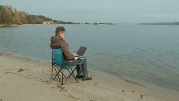 Hombre de negocios maduro sentado y relajado cerca de un río. El hombre en una chaqueta caliente y el uso de ordenador portátil . — Vídeo de stock