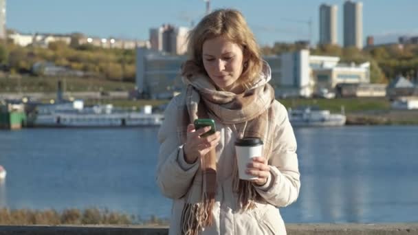 Giovane donna, in giacca bianca, nel porto fluviale, beve caffè, usa il telefono, sorride, primo piano — Video Stock