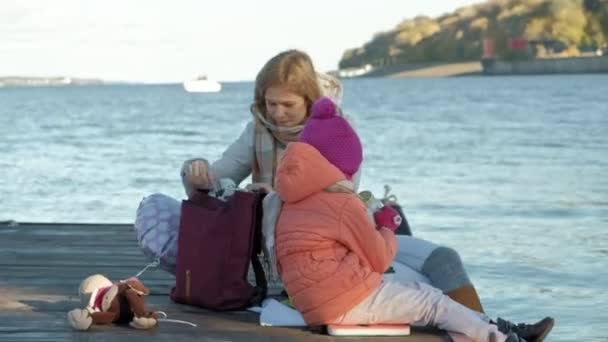 Mujer con chica, madre con hija, sentarse en el puente, río, picnic, en ropa de abrigo — Vídeo de stock