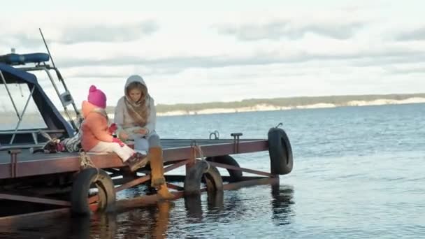 Donna con ragazza, madre con figlia, sedersi sul ponte, fiume, picnic, in abiti caldi — Video Stock