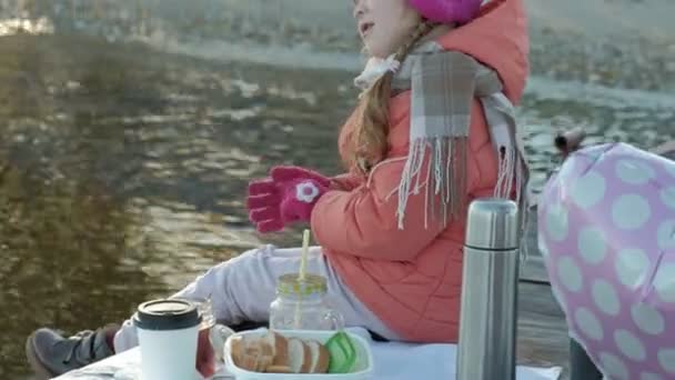 Маленька дівчинка, в теплому одязі, сидить на березі річки, на річковому порту, п'є гарячий чай, пікнік — стокове відео