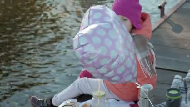 Lilla flicka, i varma kläder, sitter på stranden av floden, på flodhamnen, drycker varma te, picknick — Stockvideo