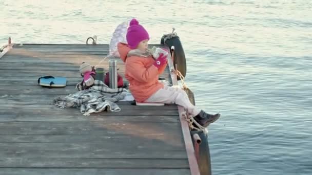 Bambina, in abiti caldi, si siede sulla riva del fiume, sul porto fluviale, beve tè caldo, picnic — Video Stock