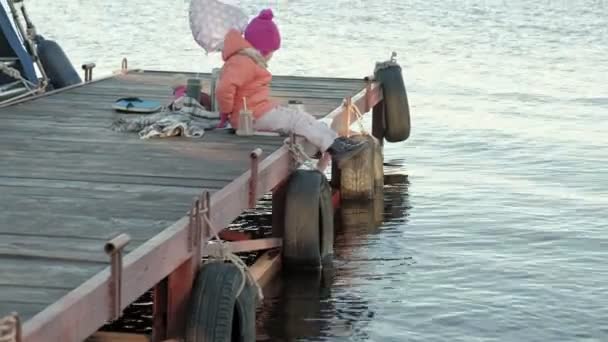 Kleines Mädchen, in warmer Kleidung, sitzt am Ufer des Flusses, am Flusshafen, trinkt heißen Tee, picknickt — Stockvideo