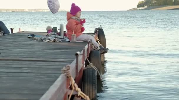 Menina, em roupas quentes, senta-se na margem do rio, no porto do rio, bebe chá quente, piquenique — Vídeo de Stock