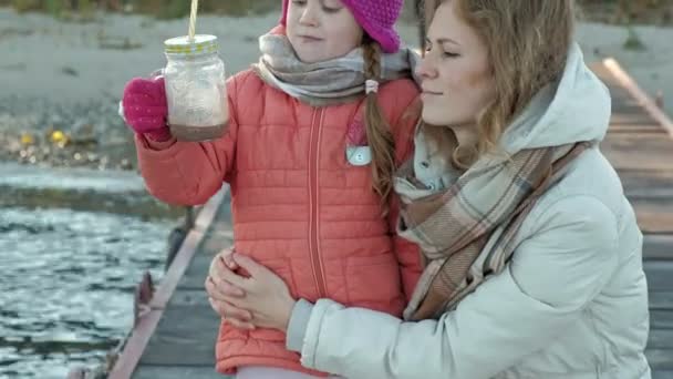 Žena s ženou, matka s dcerou, sedící na říční přístav, dívka pití kakao horké, objímání, úsměv, radost, v teplé oblečení — Stock video