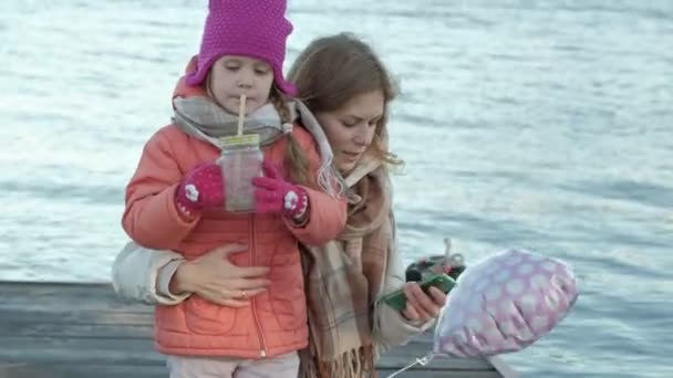 Eine Frau mit einem Mädchen, eine Mutter mit einer Tochter, die am Flusshafen sitzt, ein Mädchen, das Kakao heiß trinkt, sich umarmt, lächelnd, glücklich, in warmen Kleidern — Stockvideo