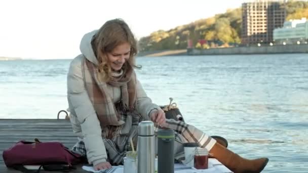 Giovane donna, in abiti caldi, si siede sulla riva del fiume, sul porto fluviale, beve tè, picnic — Video Stock