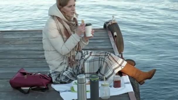 Νεαρή γυναίκα, σε ζεστά ρούχα, κάθεται στην όχθη του ποταμού, σχετικά με το λιμάνι του ποταμού, ποτά τσαγιού, πικ-νικ — Αρχείο Βίντεο