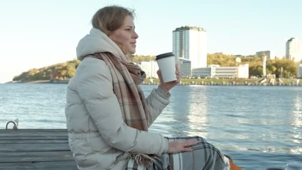 Jeune femme, en vêtements chauds, assise sur la rive de la rivière, sur le port de la rivière, boit du thé, pique-nique — Video