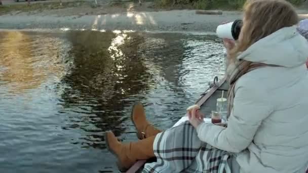 Молода жінка, в теплому одязі, сидить на березі річки, на річковому порту, п'є чай, пікнік — стокове відео