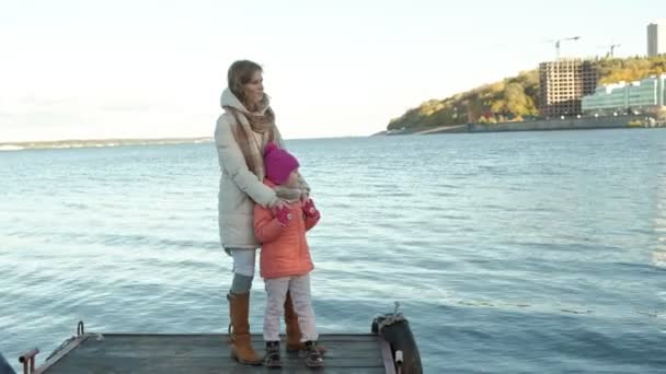 Uma mulher com uma menina, uma mãe com uma filha, estão de pé no porto do rio, de mãos dadas, abraçando, sorrindo, feliz, em roupas quentes, cobrindo-se com um tapete — Vídeo de Stock