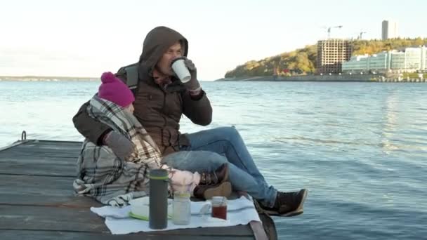 Mann und Mädchen, Vater und Tochter, sitzen im Flusshafen, trinken Tee, picknicken, lachen, warm angezogen — Stockvideo