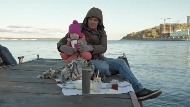 Чоловік і дівчина, батько і дочка, сидять в річковому порту, п'ють чай, пікнік, сміються, в теплому одязі — стокове відео