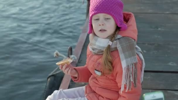 Kleines Mädchen, in warmer Kleidung, sitzt am Ufer des Flusses, am Flusshafen, trinkt heißen Tee, picknickt — Stockvideo