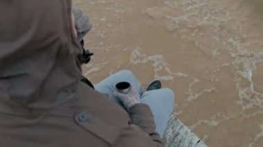 sıcak giysiler okyanus kıyısında ahşap bir bobin üzerinde oturan genç adam sıcak çay içme bir termos kullanarak bir telefon, soğuk hava, fırtına, üstten görünüm