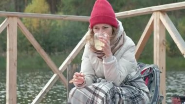 Nehir kıyısında bir halı ile kaplı bir sırt çantası ile yakın köprü üzerinde sıcak giysiler içinde bir kadın turist bir kupa sıcak çay içiyor, bir köpek aşçı gıda yakın, ishal ızgara, piknik, açık hava etkinlikleri