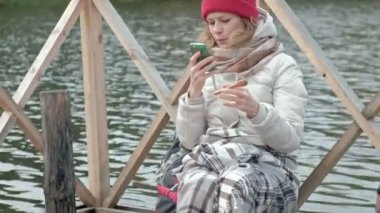 Bir kadın turist nehir kıyısında içecekler sıcak çay, bir halı ile kaplı bir sırt çantası ile yakın bir köprü üzerinde sıcak giysiler içinde bir telefon kullanan bir köpek piknik, oynayan yakın, etkin boş, sağlıklı yaşam