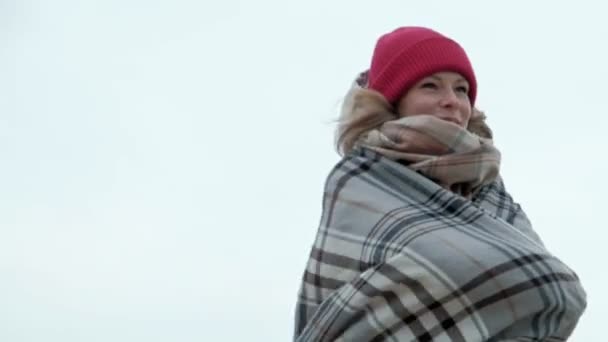 Молодая женщина в теплой одежде стоит на фоне неба, держа в руках одеяло — стоковое видео