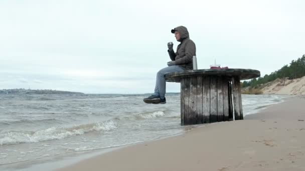 Young man in warm clothes sitting on the ocean shore, on a wooden coil, drinking hot tea from a thermos, using a telephone, cold weather, a storm — Stock Video