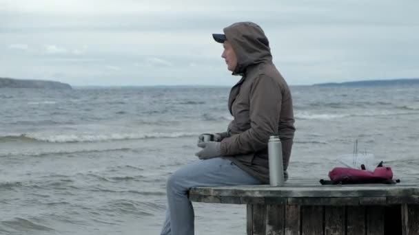 Ung man i varma kläder sitter på stranden, på en trä spole, dricka varmt te från en termos, använda en telefon, kallt väder, en storm — Stockvideo