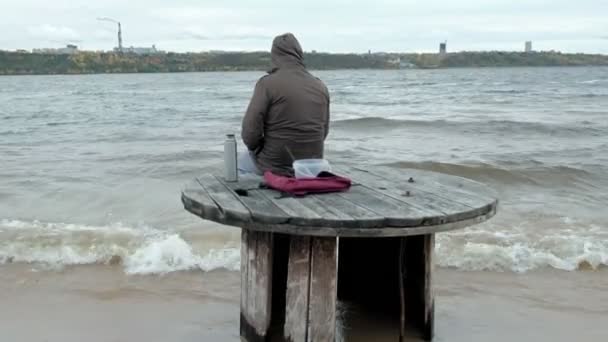年轻人在温暖的衣服坐在海边, 在一个木卷, 喝热茶从热水瓶, 使用电话, 寒冷的天气, 风暴, 后视图 — 图库视频影像