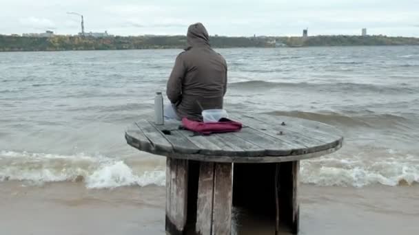 Joven en ropa de abrigo sentado en la orilla del océano, en una bobina de madera, beber té caliente de un termo, usando un teléfono, clima frío, tormenta, vista trasera — Vídeos de Stock