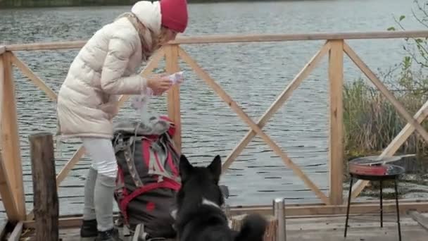 Eine Touristin sitzt auf der Brücke in der Nähe des Flussufers, füttert den Hund, kocht Futter, Picknick, aktive Erholung und einen gesunden Lebensstil im Freien. Reisekonzept — Stockvideo