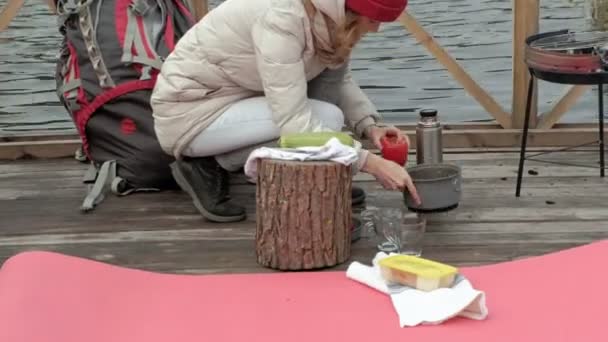 Turysta Kobieta w ciepłe ubrania na moście w pobliżu brzegu rzeki, z plecakiem, pies gra u boku, gotuje jedzenie na grilla, warzywa myje, piknik, na świeżym powietrzu i zdrowego stylu życia — Wideo stockowe