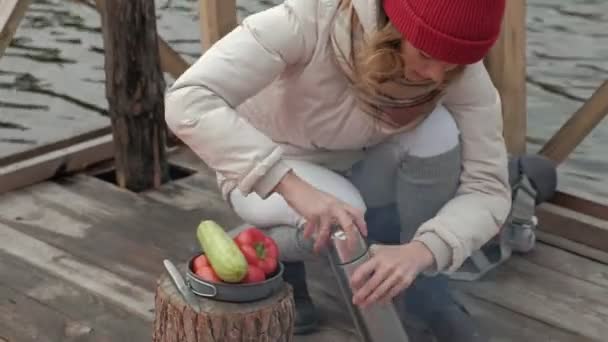 Žena turista v teplé oblečení na mostě poblíž břehu řeky s batohem, pes hraje po boku, vaří jídlo na grilu, mytí zeleniny, piknik, Outdoorové aktivity a zdravého životního stylu — Stock video