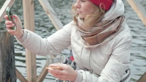 Uma turista mulher em roupas quentes em uma ponte perto da margem do rio com uma mochila, coberta com um tapete, bebe chá quente, usa um telefone, cozinha comida na grelha, piquenique, um cão brinca nas proximidades, ativo — Vídeo de Stock