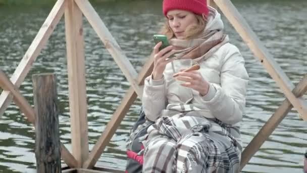 Женщина турист в теплой одежде на мосту у берега реки с рюкзаком, покрытый ковром, пьет горячий чай, пользуется телефоном, пикник, собака играет поблизости, активный досуг, здоровый образ жизни в — стоковое видео