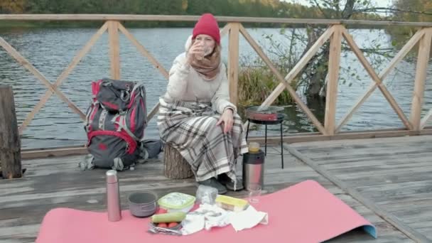 Una turista en ropa de abrigo en un puente junto a la orilla del río con una mochila, mira a su alrededor, se refugia en una alfombra, bebe té caliente, usa el teléfono, cocina comida en la parrilla, picnic, actividades al aire libre — Vídeo de stock