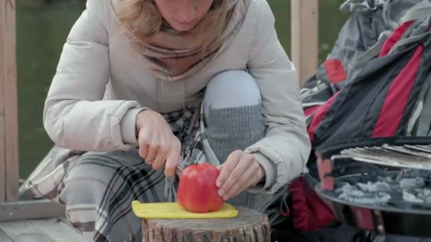 Turista mujer en ropa de abrigo en un puente cerca de la orilla del río con una mochila, tamiza verduras, preparando comida en la parrilla, picnic, descanso activo, estilo de vida saludable. Concepto de viaje — Vídeos de Stock