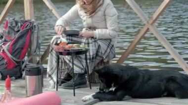 Kadın turist nehir kıyısında bir sırt çantası ile yakın köprü üzerinde sıcak giysiler içinde ızgara sebzeler hazırlar, beslemek köpek, piknik, açık hava etkinlikleri, sağlıklı bir yaşam tarzı. Seyahat kavramı
