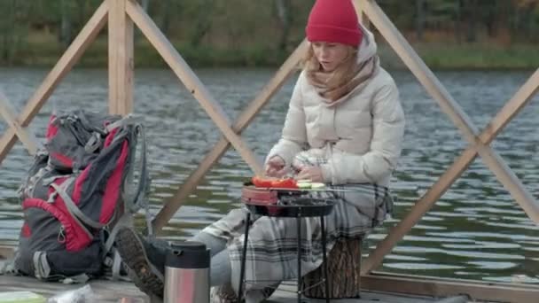 Vrouw toerist in warme kleren op de brug in de buurt van de bank van de rivier met een rugzak, bereidt gegrilde groenten, een picknick, actieve rust, gezonde levensstijl. Travel concept — Stockvideo