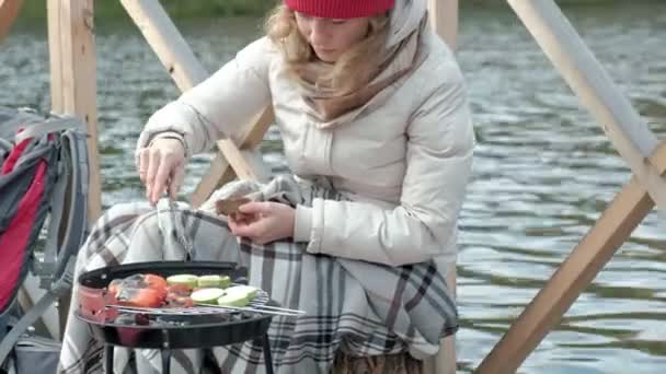 Touristin in warmer Kleidung auf der Brücke am Flussufer mit Rucksack, bereitet gegrilltes Gemüse zu, füttert den Hund, Picknick, Outdoor-Aktivitäten, gesunder Lebensstil. Reisekonzept, Nahaufnahme — Stockvideo