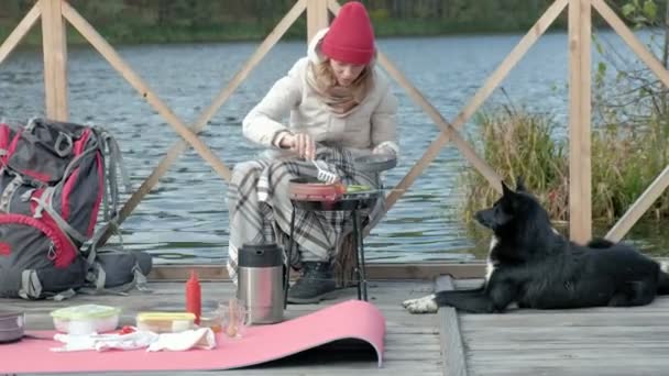 Donna turista in abiti caldi sul ponte vicino alla riva del fiume con uno zaino, prepara verdure alla griglia, alimenta il cane, pic-nic, attività all'aperto, stile di vita sano. Concetto di viaggio — Video Stock